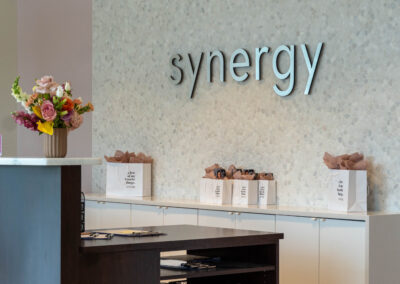 Polished silver 'Synergy' sign on a mosaic stone wall at the reception, with decorative flowers and branded bags to the side."
