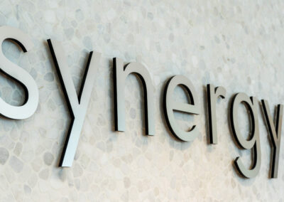 "Metallic 'Synergy' sign over a peaceful lobby backdrop, accented by a floral arrangement and customized company shopping bags."