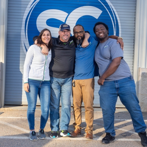 "Four diverse and cheerful employees of SignCraft Solutions standing together outside their office, posing for a group photo with the company's logo prominently displayed on the building behind them."