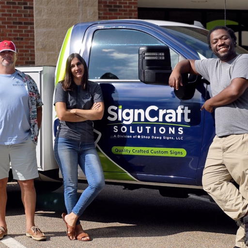" three diverse and cheerful employees of SignCraft Solutions standing together outside their office, posing for a group photo with the company's logo prominently displayed on the building behind them."